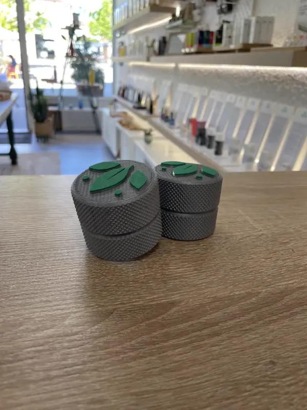 Deux petites boîtes grises moletées au couvercle à logo vert, imprimées en 3D et posées sur le comptoir d'une boutique