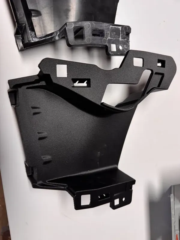 Pièce technique noire de carrosserie automobile, refaite sur mesure en impression 3D, présentée contre un mur blanc