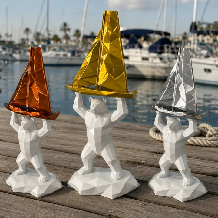 Trois trophées imprimés en 3D posés sur un ponton : un ours blanc à facettes soulève un voilier bronze, or ou argent
